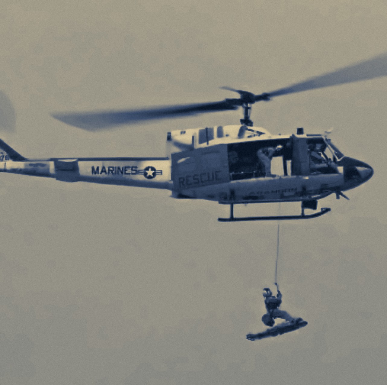 A search and rescue corpsman on the hoist line beneath a Marines rescue helicopter, with a rescue litter